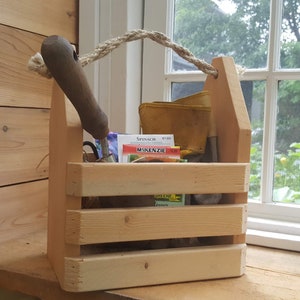 May include: A wooden gardening caddy with a rope handle. The caddy is filled with gardening tools, seed packets, and other gardening supplies. The caddy is sitting on a wooden surface next to a window.