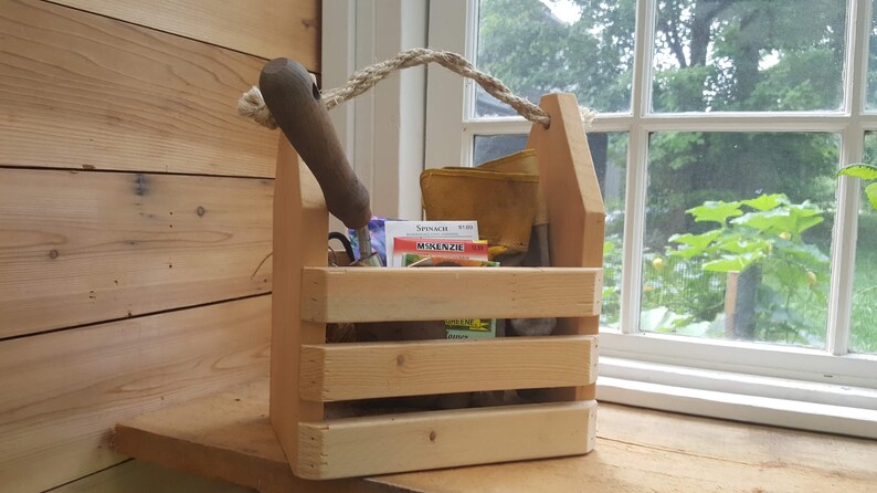 May include: A wooden gardening caddy with a rope handle. The caddy is filled with gardening tools, seed packets, and other gardening supplies. The caddy is sitting on a wooden surface next to a window.