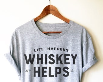 Könnte beinhalten: Ein hellgraues T-Shirt mit dem schwarzen Aufdruck "LIFE HAPPENS WHISKEY HELPS". Das Shirt hängt an einem Holzkleiderbügel. Die Ärmel sind hochgekrempelt.