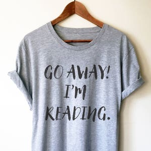 Puede incluir: Una camiseta gris con el texto "GO AWAY! I'M READING." impreso en negro.
