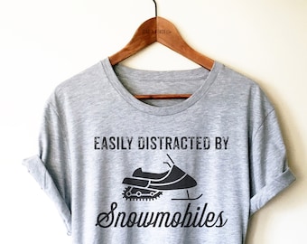 Puede incluir: Camiseta gris con un gráfico negro de una moto de nieve y el texto "Easily Distracted By Snowmobiles".