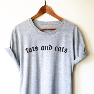 May include: A heather grey t-shirt with the text "tats and cats" printed in black gothic font.