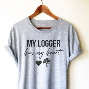 Op de afbeelding: Een heidekleurig t-shirt met de tekst "MY LOGGER has my heart" in zwarte letters. Onder de tekst staan een zwart hart en een zwarte boom.
