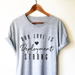 May include: Grey t-shirt with black text that reads "Our love is Deployment Strong" with a small black heart above the word "Deployment".