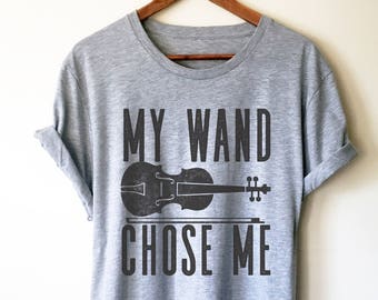 May include: A light grey t-shirt featuring the text "MY WAND CHOSE ME" in black, accompanied by a violin graphic. The garment is displayed on a wooden hanger against a white backdrop.