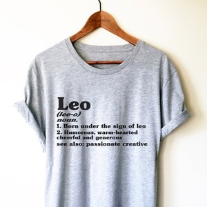 May include: Gray t-shirt with black text that reads "Leo (lee-o) noun. 1. Born under the sign of leo 2. Humorous, warm-hearted cheerful and generous see also: passionate creative"