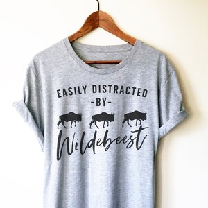 Może przedstawiać: Szary T-shirt z napisem "EASILY DISTRACTED -BY- Wildebeest" i trzema czarnymi sylwetkami gnu.