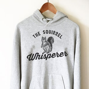 May include: A gray hooded sweatshirt with the text "The Squirrel Whisperer" printed on the front. The text is in black and features a black and white illustration of a squirrel.