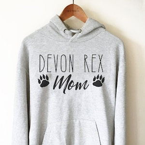 May include: A light gray hoodie with the text "DEVON REX Mom" in black, along with paw print graphics. The hoodie has a drawstring hood and a front pocket. The garment is hanging on a wooden hanger.