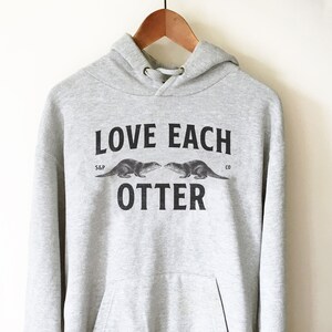 Peut inclure: Un sweat &agrave; capuche gris avec le texte "LOVE EACH OTTER" imprim&eacute; sur le devant. Le texte est en noir et il y a deux illustrations de loutres au-dessus du texte.
