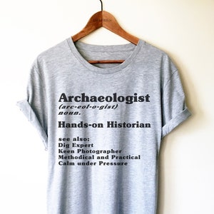Op de afbeelding: Grijs T-shirt met zwarte tekst die luidt "Archeoloog (arc-eol-o-gist) zelfstandig naamwoord. Hands-on historicus Zie ook: Graafexpert Gezellige fotograaf Methodisch en praktisch Rustig onder druk"