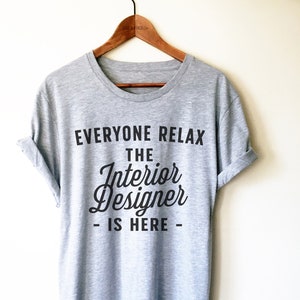 Op de afbeelding: Grijs T-shirt met zwarte tekst die "Everyone Relax The Interior Designer - Is Here -" luidt.