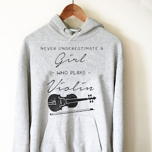 May include: Gray hooded sweatshirt with a black graphic print that says "Never underestimate a girl who plays violin" with a black silhouette of a violin and bow.