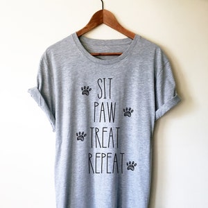 May include: A heather gray t-shirt with the words "SIT PAW TREAT REPEAT" in black, along with paw print graphics. The shirt is hanging on a wooden hanger.
