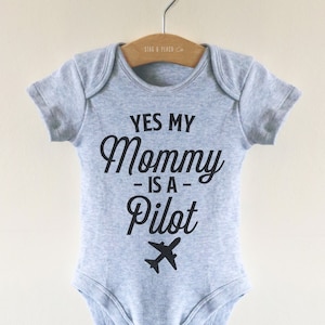 Può includere: Un body per neonato grigio con il testo "Yes My Mommy Is A Pilot" e un piccolo aereo grafico.
