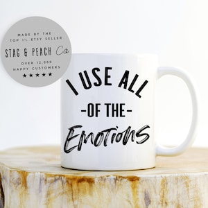 Puede incluir: Taza de café de cerámica blanca con texto negro que dice "Uso todas las emociones".