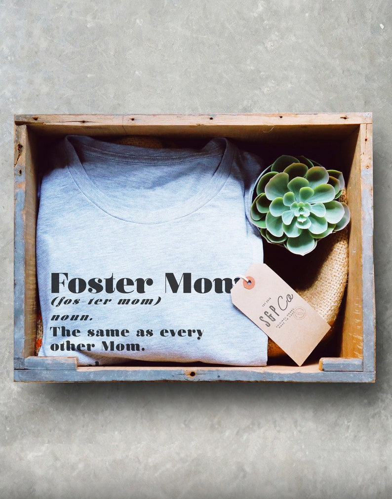 Pu&ograve; includere: Una maglietta grigia con la scritta "Foster Mom" e una definizione, presentata in una cassa di legno con una succulenta e un'etichetta. L'etichetta recita "S&P Co."