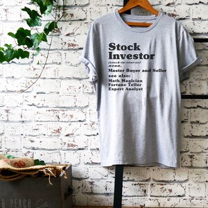 Op de afbeelding: Een grijs T-shirt met de tekst "Stock Investor (stock-in-vest-or) noun. Master Buyer and Seller see also: Math Magician Fortune Teller Expert Analyst" gedrukt op de voorkant.