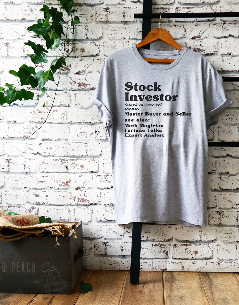Op de afbeelding: Een grijs T-shirt met de tekst "Stock Investor (stock-in-vest-or) noun. Master Buyer and Seller see also: Math Magician Fortune Teller Expert Analyst" gedrukt op de voorkant.