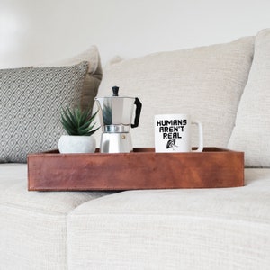 Puede incluir: Una taza de caf&eacute; blanca con el texto "HUMANS AREN'T REAL" y una ilustraci&oacute;n en blanco y negro de un OVNI. La taza est&aacute; sobre una bandeja de cuero marr&oacute;n con una cafetera plateada y una peque&ntilde;a planta en maceta.