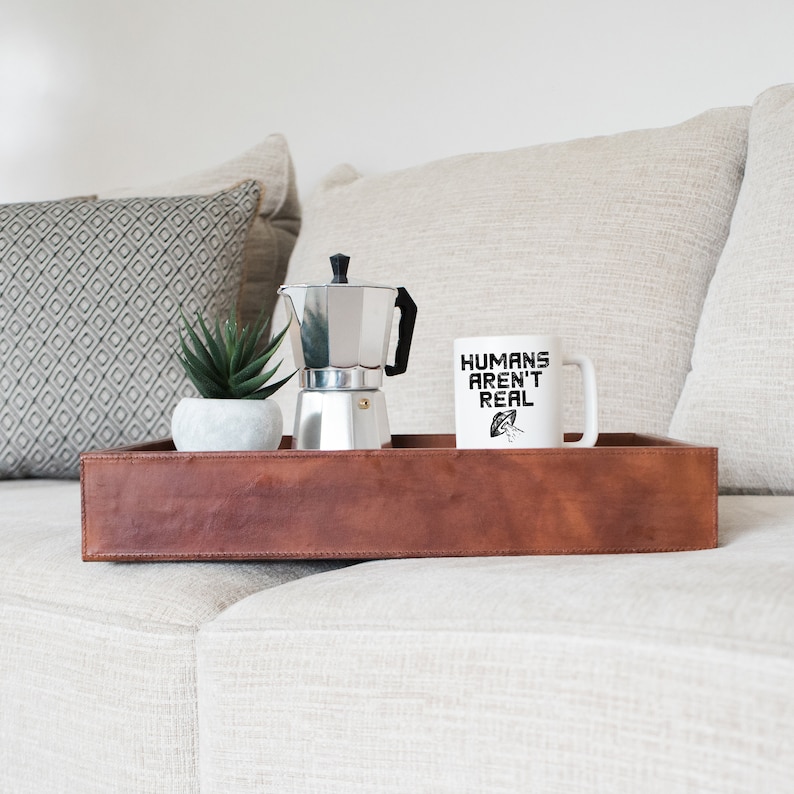 Puede incluir: Una taza de caf&eacute; blanca con el texto "HUMANS AREN'T REAL" y una ilustraci&oacute;n en blanco y negro de un OVNI. La taza est&aacute; sobre una bandeja de cuero marr&oacute;n con una cafetera plateada y una peque&ntilde;a planta en maceta.