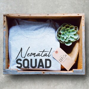 May include: A gray t-shirt with the text "Neonatal Squad" in black lettering. The t-shirt is in a wooden crate with a small succulent plant and a brown tag with the text "S&P Co."