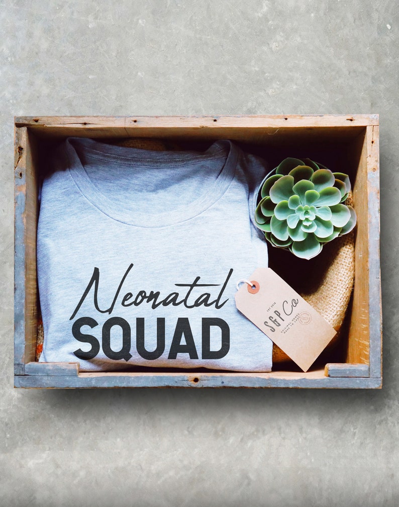 May include: A gray t-shirt with the text "Neonatal Squad" in black lettering. The t-shirt is in a wooden crate with a small succulent plant and a brown tag with the text "S&P Co."