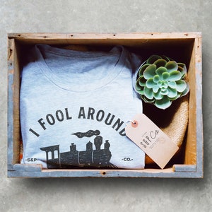 May include: A gray t-shirt with a black and white graphic of a train and the text "I Fool Around -S&P- Co."  The t-shirt is in a wooden crate with a small green succulent plant and a brown tag with the text "S&P Co."
