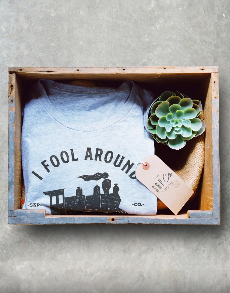 May include: A gray t-shirt with a black and white graphic of a train and the text "I Fool Around -S&P- Co."  The t-shirt is in a wooden crate with a small green succulent plant and a brown tag with the text "S&P Co."