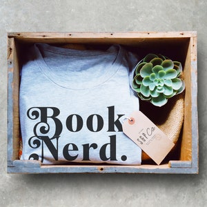 May include: A gray t-shirt with the text "Book Nerd." in black lettering. The t-shirt is in a wooden crate with a small succulent plant and a brown tag with the text "SFF Co." in black lettering.