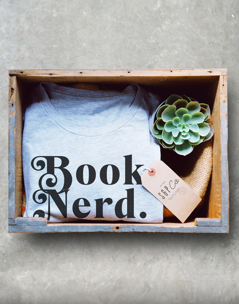 May include: A gray t-shirt with the text "Book Nerd." in black lettering. The t-shirt is in a wooden crate with a small succulent plant and a brown tag with the text "SFF Co." in black lettering.