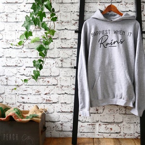 Puede incluir: Sudadera gris con capucha con el texto "Happiest When It Rains" en letras de escritura negra.
