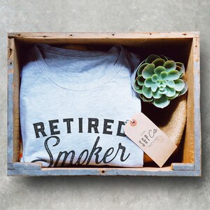 May include: A gray t-shirt with the text "Retired Smoker" in black lettering. The shirt is in a wooden crate with a small succulent plant and a brown tag that says "SGP Co."