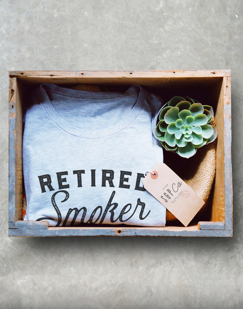 May include: A gray t-shirt with the text "Retired Smoker" in black lettering. The shirt is in a wooden crate with a small succulent plant and a brown tag that says "SGP Co."