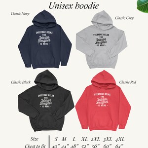Op de afbeelding: Unisex hoodie in vier kleuren: marine, zwart, grijs en rood. De hoodie heeft de tekst "Everyone Relax The Interior Designer - Is Here -" in witte letters. De maattabel toont maten S tot en met 4XL met bijbehorende borstmaten in inches.