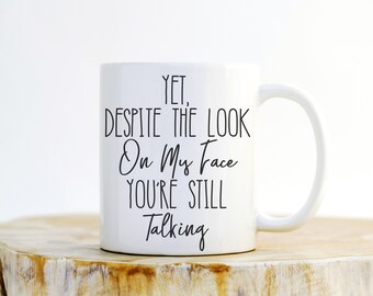 May include: White ceramic mug with black text that reads "Yet, despite the look on my face you're still talking".