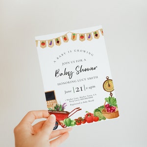 May include: A white baby shower invitation with a watercolor illustration of vegetables and a wheelbarrow. The text reads "A baby is growing. Join us for a baby shower honoring Lucy Smith. June 21, 2-4pm. Market Lane, Market Town, CA. Rsvp Lacey 888.888.8888. Registered at Baby World."