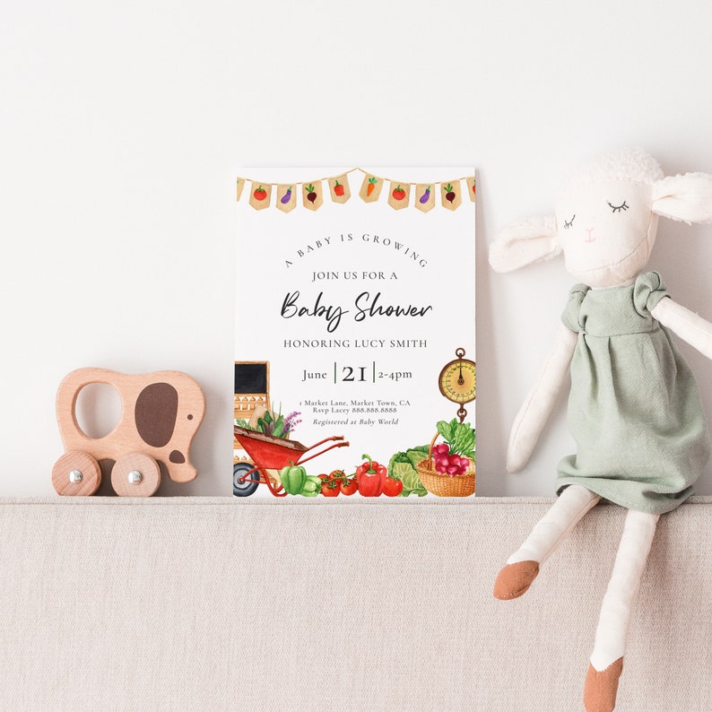 May include: A baby shower invitation with a watercolor illustration of a red wagon filled with fresh produce. The invitation reads "A baby is growing. Join us for a baby shower honoring Lucy Smith. June 21, 2-4pm. Market Lane, Market Town, CA. RSVP Lucy 555-555-5555. Registered at Baby World."