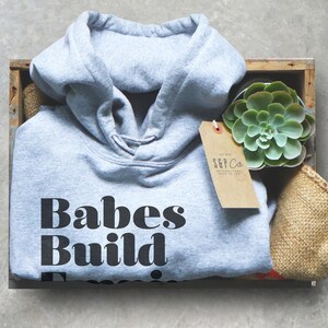 May include: A gray hoodie with the text "Babes Build Empires" in a bold, black font. The hoodie is folded inside a wooden crate, next to a succulent plant and a tag that reads "S&P Co."