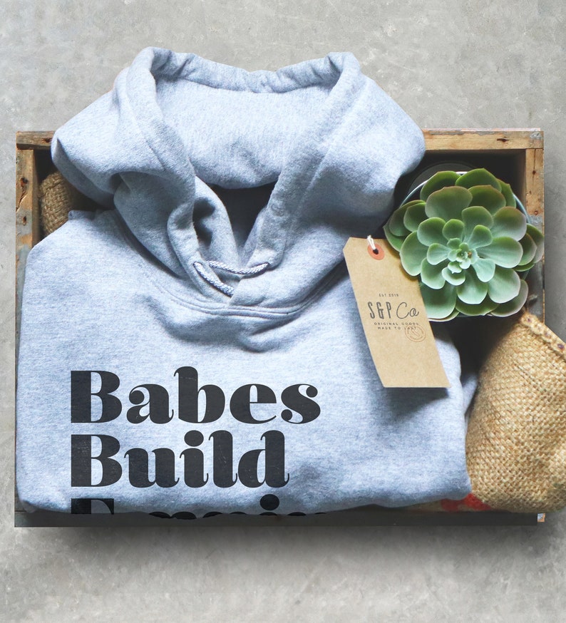 May include: A gray hoodie with the text "Babes Build Empires" in a bold, black font. The hoodie is folded inside a wooden crate, next to a succulent plant and a tag that reads "S&P Co."