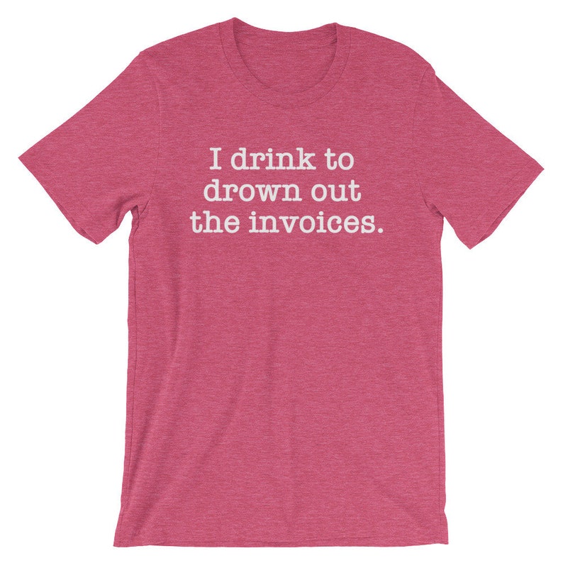 Puede incluir: Una camiseta de color rosa claro con el texto "I drink to drown out the invoices." impreso en blanco.