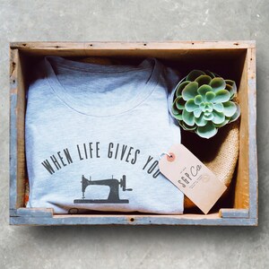 May include: A gray t-shirt with the text "When life gives you..." and a vintage sewing machine graphic. The shirt is in a wooden crate with a succulent plant and a brown tag with the text "SGP Co."