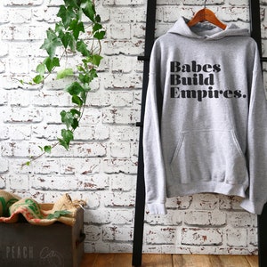 May include: A gray hoodie with the text "Babes Build Empires" in black, hanging on a wooden hanger. The hoodie has a front pocket and a hood. The background features a white brick wall and a black ladder.