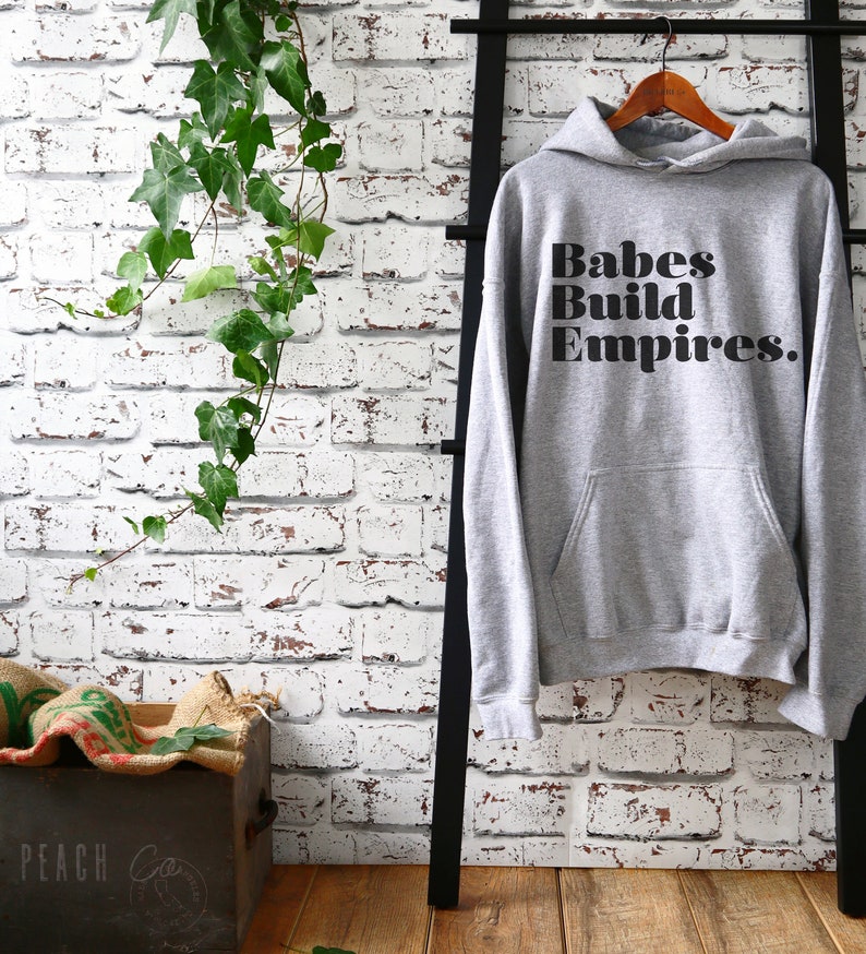 May include: A gray hoodie with the text "Babes Build Empires" in black, hanging on a wooden hanger. The hoodie has a front pocket and a hood. The background features a white brick wall and a black ladder.