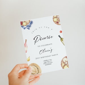 May include: A white invitation card with a watercolor illustration of a picnic basket with food and wine. The text on the card reads "Join us for a Picnic to celebrate Olivia's 21st Birthday Party". The invitation includes the address and RSVP information.