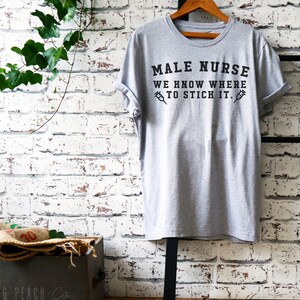 Puede incluir: Una camiseta gris con el texto "MALE NURSE WE KNOW WHERE TO STICK IT." impreso en negro. El texto est&aacute; rodeado por dos s&iacute;mbolos de jeringas negras.