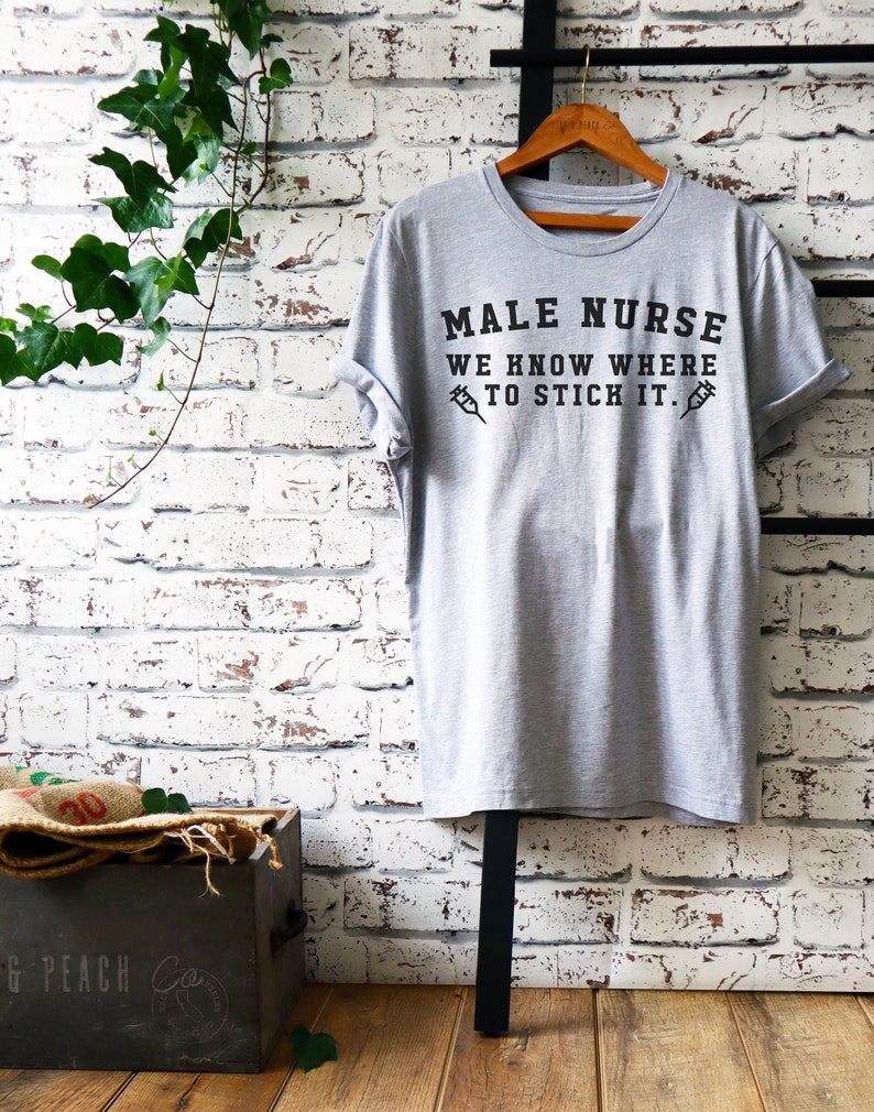 Puede incluir: Una camiseta gris con el texto "MALE NURSE WE KNOW WHERE TO STICK IT." impreso en negro. El texto est&aacute; rodeado por dos s&iacute;mbolos de jeringas negras.