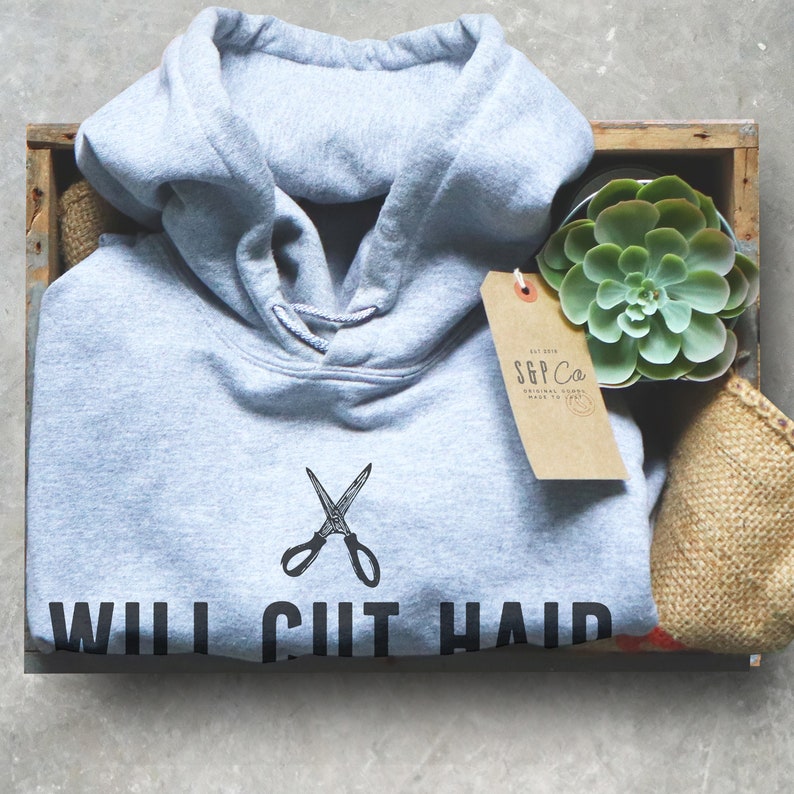 Op de afbeelding: Een lichtgrijze hoodie met een zwarte afbeelding van een schaar en de tekst "Will I Cut Hair". De hoodie bevindt zich in een houten doos met een kleine potplant met een vetplant en een bruin label met de tekst "SGP Co."