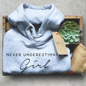 May include: A light gray hooded sweatshirt with the text "NEVER UNDERESTIMATE A Girl" in black lettering. The sweatshirt is in a wooden crate with a small potted succulent plant and a brown paper tag with the text "S&P Co. ORIGINAL GOODS MADE TO LAST" in black lettering.