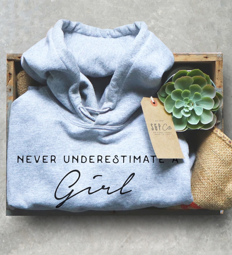May include: A light gray hooded sweatshirt with the text "NEVER UNDERESTIMATE A Girl" in black lettering. The sweatshirt is in a wooden crate with a small potted succulent plant and a brown paper tag with the text "S&P Co. ORIGINAL GOODS MADE TO LAST" in black lettering.